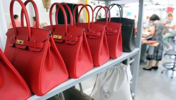 Six Hermès Birkin bags (five red, one black) are lined up next to each other on a table.