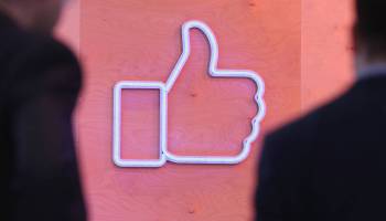 People walk past a large sign of the Facebook 'Like' symbol.