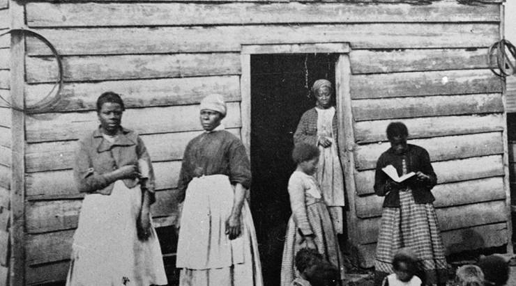A group of women and children, presumably slaves, sit and stand around the doorway of a rough wooden cabin in the southern United States in the mid-19th century. One girl reads a book to the group of sitting children.