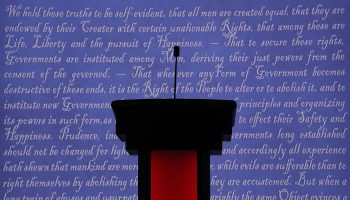 A red and black debate lectern with a slim microphone against a stage background featuring the words to the declaration of independence.