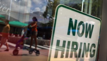A "now hiring" sign is seen in a storefront window.