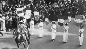 In an archival black and white photo, women dressed in white march while holding signs representing various groups in support of womens' suffrage.