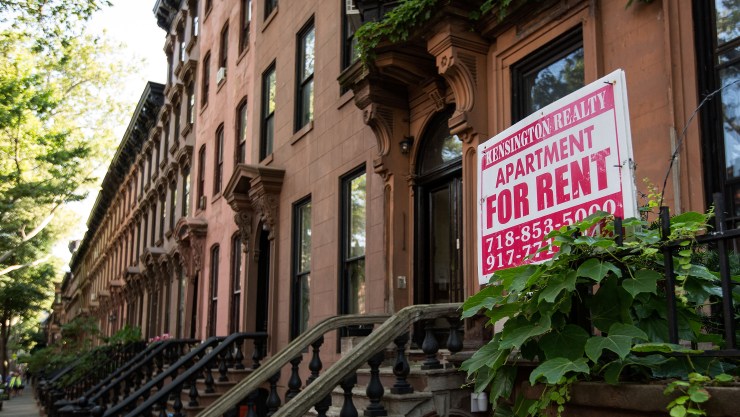 A sign advertises an apartment for rent along a row of brownstone townhouses in New York City.