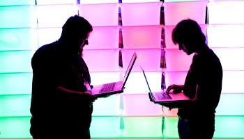 Two people use their laptops in front of a light wall.