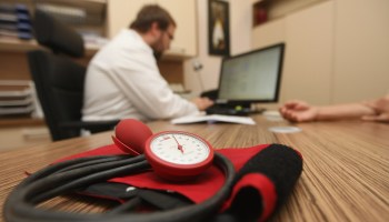 A blood pressure cuff lies on a desk as a doctor works on a computer across from a patient.