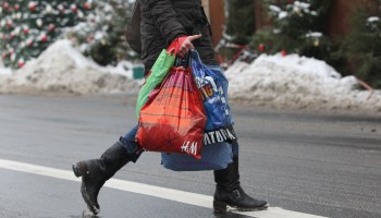 A person dressed in winter clothing crosses a snow-lined street while holding shopping bags.