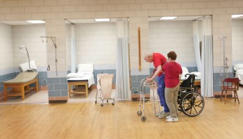 A health care aide helps an elderly man who is using a walker in a medical facility.