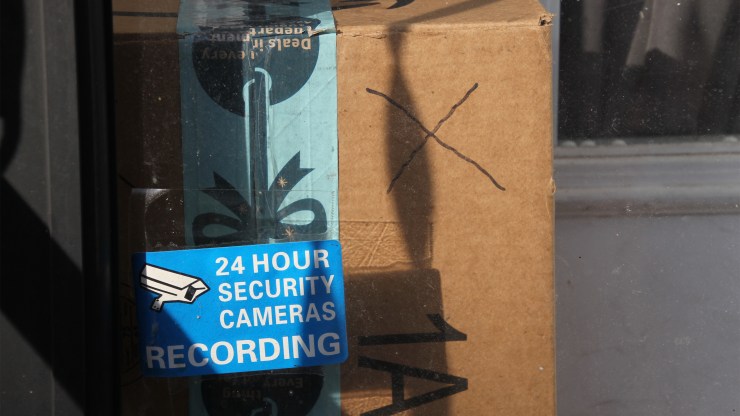 A package on a porch with a 24 hour security sign