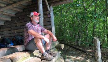 Kevin Rosenberg sits on the steps of a log shed in the woods