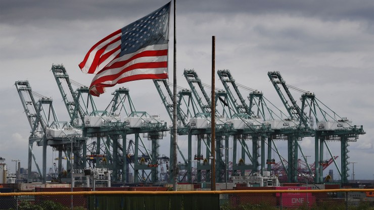 The US flag flies in a gray sky over shipping cranes and containers in Long Beach, California on March 4, 2019.