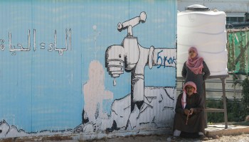 A woman stands behind a sitting man and a mural on a temporary shelter showing a dripping tap with a caption reading in Arabic "Water = Life" at the Za'atari refugee camp in northern Jordan in July.