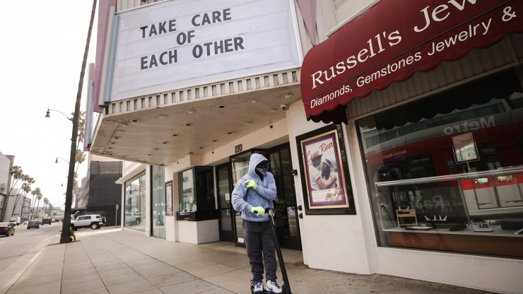 A man wears gloves and a bandanna across his face while riding a scooter past a shuttered movie theater, with the message Take Care of Each Other displayed on the marquee, on March 18