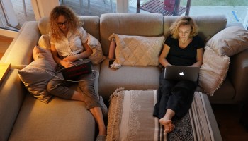 A mother and daughter in loungewear work on their laptops on the couch.
