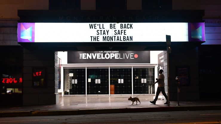 A man walks his dog past a closed theater with "We'll Be Back, Stay Safe" on the marquee.