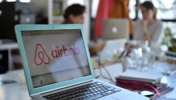 Airbnb on a laptop screen with two people in the background.