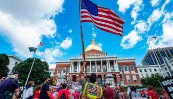 A man with a neon yellow QAnon vest holds up an American flag outside the Massachusetts State House as part of a protest against vaccines.