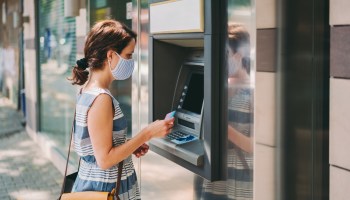 Woman with protective face mask is using credit card to withdraw money at the ATM