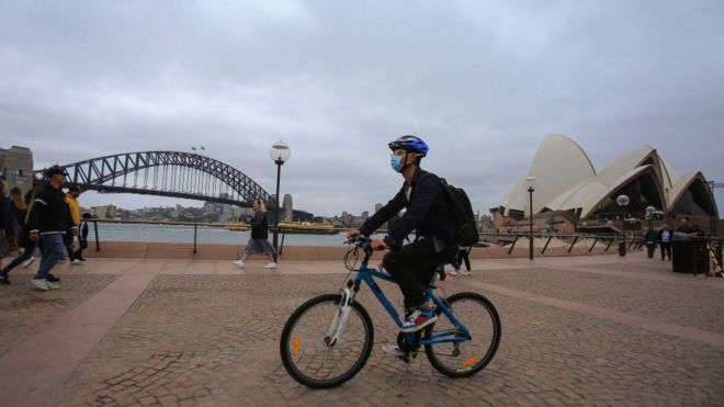 A man in a face mask cycles close to Sydney Sydney Harbour Bridge and Opera House.