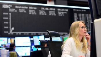 A trader sits at her desk under the day's performance board.