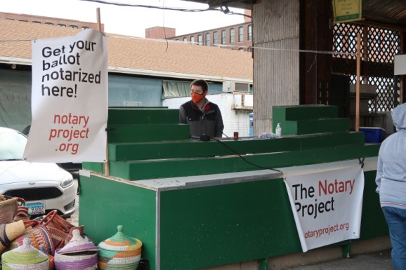 Will Stevens stands behind a green wooden barrier usually used to display goods for sale at the Soulard Farmers Market in St. Louis. The mostly empty display hold just his notary book and stamp, a bag of pens, and a bottle of hand sanitizer. On the front of the booth and to the side hang large white banners reading "The Notary Project" and "Get Your Ballot Notarized Here!"