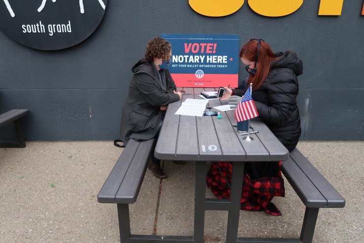 Jill Anderson sits at a gray picnic table with Cynthia Barounis as Anderson notarizes Barounis' ballot envelop. On the table is a large sign reding "Vote! Notary Here". In the foreground, a small American flag sits at the front of the table.