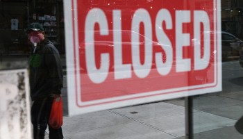 A person wearing a face mask is reflected in the window of a closed business, next to a CLOSED sign in New York City.