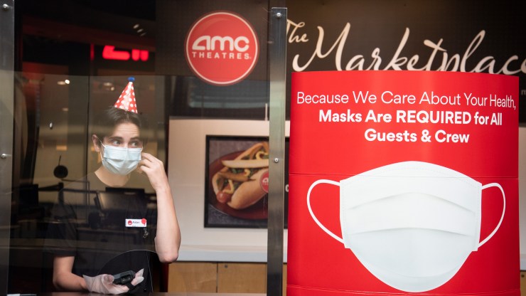 A worker wearing a face mask, gloves and party hat at stands next to a sign about face mask requirements a reopened AMC theatre in Highlands Ranch, Colorado, in August.