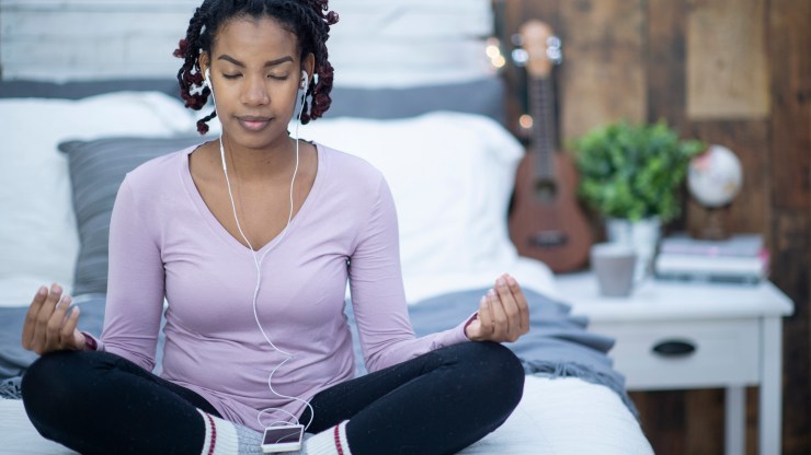 A woman sits on her bed cross-legged with her hands on her knees, meditating while listening to something on her phone.