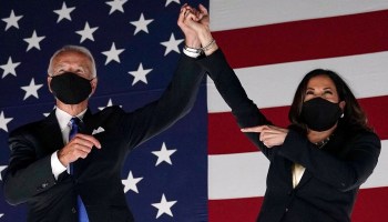 Joe Biden and Kamala Harris, wearing masks, grab hands in front of an American flag at a campaign event in August.