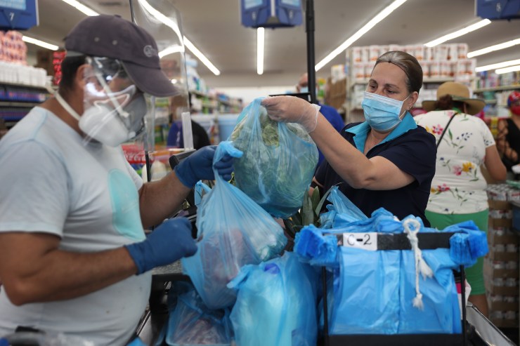 A grocery store worker stands behind a partial protective plastic screen and wears a mask and gloves as she works as a cashier.