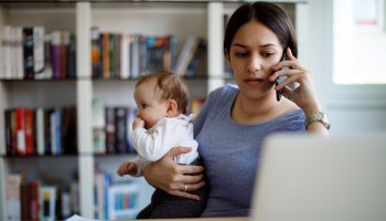A worried mom holds her baby in one arm and holds a phone up to her ear with her other hand, while looking at her laptop.