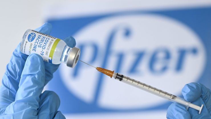 A photo illustration showing a vaccine syringe and vial, with the logo of Pfizer in the background.