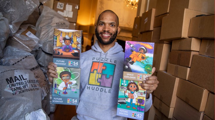 Puzzle Huddle founder Matthew Goins holding up four of his puzzles in his family’s living room, which currently doubles as a warehouse.
