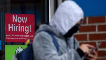 A man wearing a face mask stands next to a "Now Hiring " sign in front of a store on Dec. 18, 2020, in Arlington, Virginia.