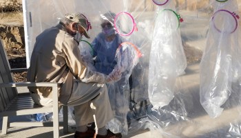 A man holds his wife's hands through the wall of a plastic "hug tent" outside a Colorado nursing facility. Elderly people in long-term care are prioritized for COVID-19 vaccination.