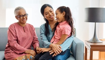 A smiling multigenerational family, comprised of a grandmother, mother and daughter, sits on a sofa.
