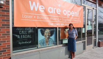 Shobha Tummala stands in front of her "Shobha" location in Rockville, MD. Stretched across the glass windows of the salon is a large orange sign that reads "We are open!"