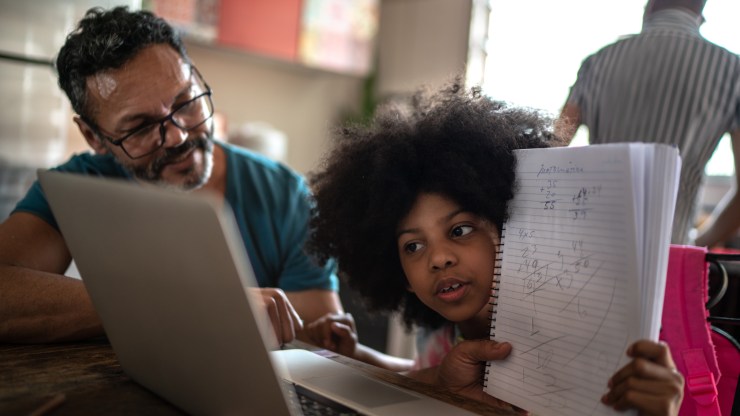 A father helps his daughter share her schoolwork via a video call at their kitchen table.