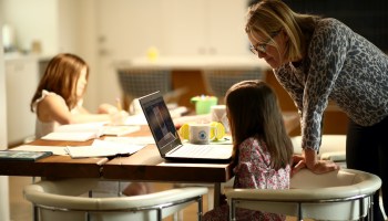 A woman helps her two children with schoolwork from home via laptop.