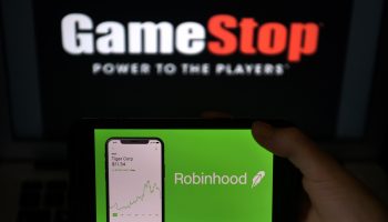 A person holds a screen up displaying the Robinhood app with a GameStop sign in the background.