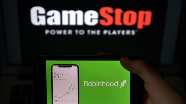 A person holds a screen up displaying the Robinhood app with a GameStop sign in the background.