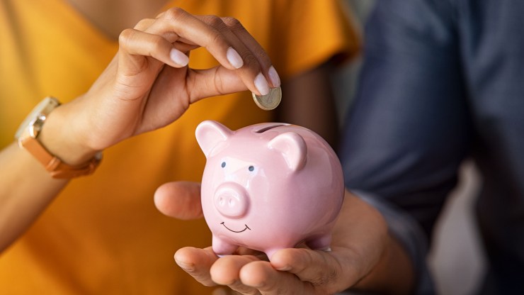 This photo illustration shows someone holding a pink piggy bank while another person puts a coin in it.