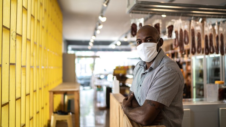A Black small business owner wearing a face mask stands near the entrance of his business.