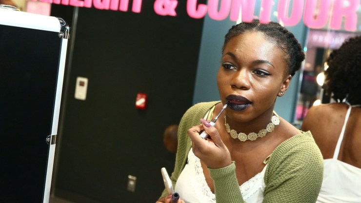 A woman applies matte lipstick in a mirror.