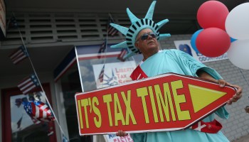 A man wearing a Statue of Liberty costume holds a sign reading, "It's tax time."