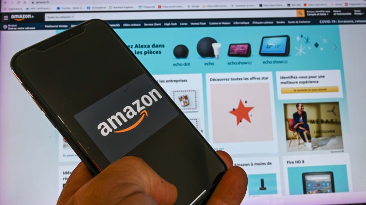 In this photograph taken on November 18, 2020, a person poses with a smartphone showing an Amazon logo, in front of a computer screen displaying the home page of Amazon.