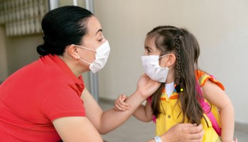 A mother bends down to speak with her daughter on the first day of school. They're both wearing face masks.