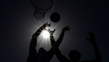 Two basketball players jump and reach for a loose ball. You can see the silhouettes of the players and a hoop against the sun.