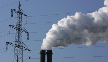 An electricity pylon stands in front of the exhaust pluming from the main chimneys of a coal-fired power plant.