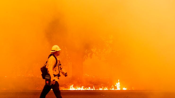 A firefighter walks down as a road as a fire burns on, turning the air orange.